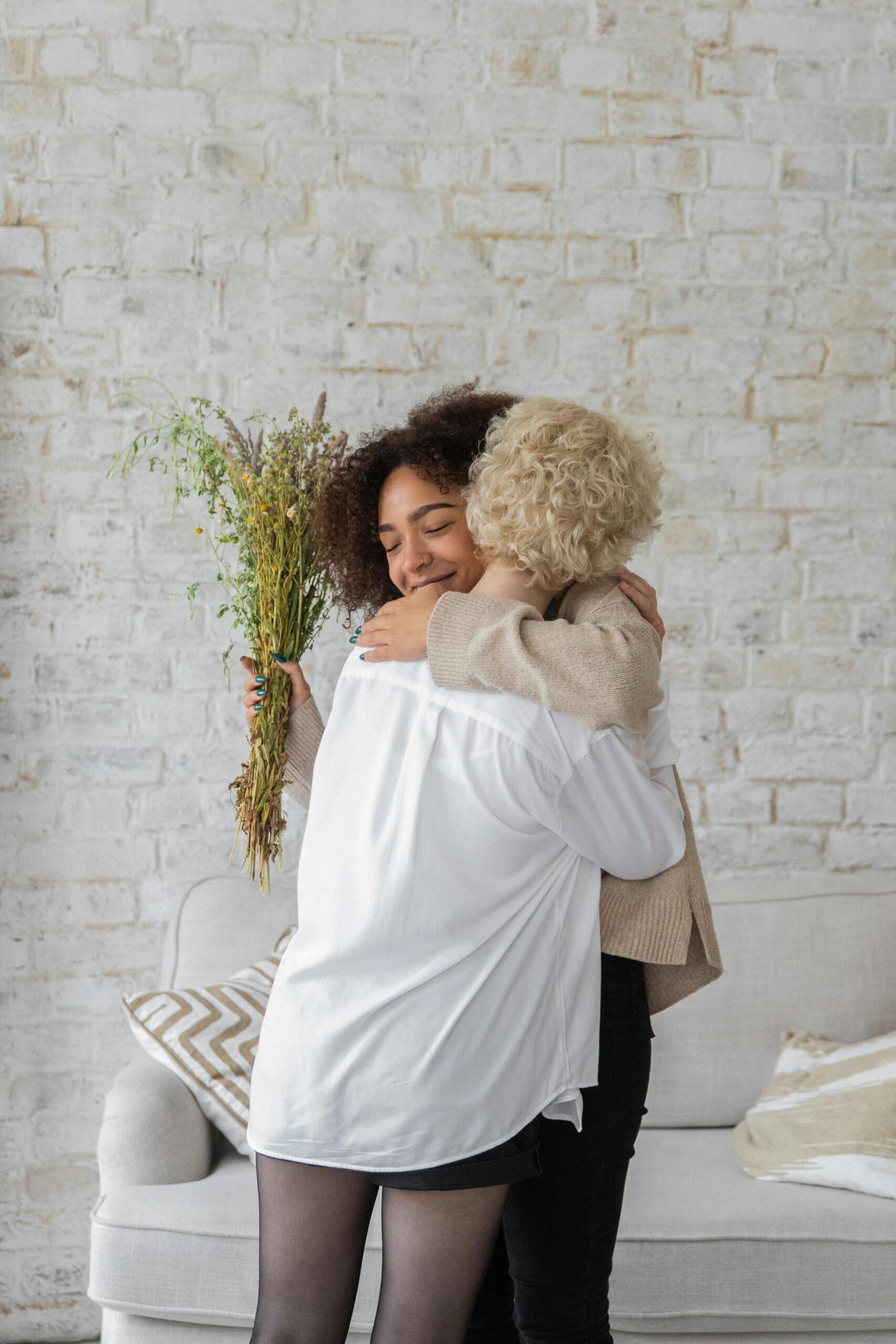 African American lady with bouquet of flowers embracing anonymous female friend while standing in room near couch against brick wall